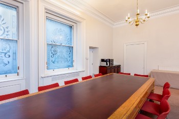 white room with long table surrounded by red chairs and two sash windows